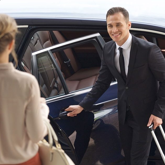A chauffeur in a suit stands by a luxury car, holding a door open for a passenger for airport shuttle service